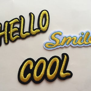 May include: Three embroidered patches with the words "HELLO", "Smile", and "COOL". The patches feature yellow text with black outlines. The "Smile" patch has a light blue border.