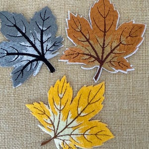 Puede incluir: Tres parches bordados con forma de hoja de arce y costuras detalladas. Un parche es gris con venas negras, otro es naranja y marrón con venas oscuras, y el tercero es amarillo y crema con venas marrones. Cada hoja tiene un borde blanco.