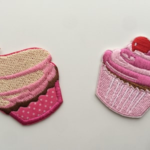 May include: Two embroidered cupcake patches. One is pink with white frosting and a red cherry on top. The other is pink and white with brown frosting and white polka dots.