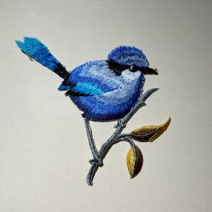 Puede incluir: Parche bordado de un pájaro azul con pico negro, posado en una rama con dos hojas de color marrón dorado. Las plumas del pájaro están detalladas con diferentes tonos de azul. La rama es gris.