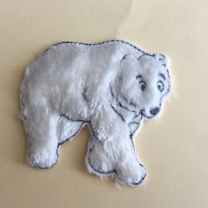 May include: A white plush polar bear applique with black embroidered details. The bear is walking with its head turned to the side.