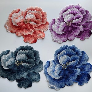 May include: Four embroidered fabric flower patches in different colors. The patches are in shades of pink, purple, blue, and gray. The flowers are all the same design, with a white center and multiple layers of petals.