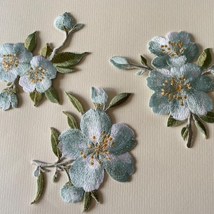May include: Embroidered floral appliques in shades of blue, white, and green. The flower patches feature detailed stitching with yellow centres and green leaves. These decorative patches are ideal for fabric crafts.