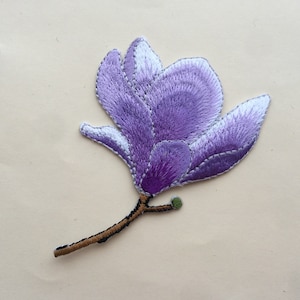 May include: An embroidered flower patch in purple, with a brown stem and a green bud. The flower is a magnolia blossom with a white centre.
