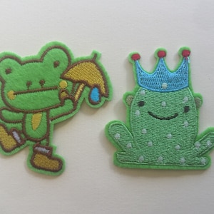 May include: Two green frog embroidered patches. One frog is holding a yellow umbrella and the other frog is wearing a blue crown.