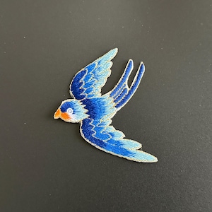 May include: An embroidered patch in the shape of a bird in flight. The bird features shades of dark and light blue thread with a bright orange beak and a white eye detail. The edges of the wings and tail are outlined in a metallic gold thread.