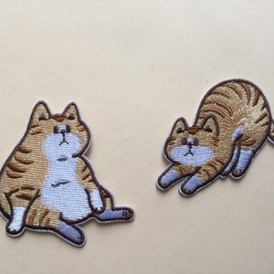 May include: Two embroidered iron-on patches featuring cartoon cats. One cat is sitting with its paws crossed and the other is stretching. Both cats are brown and white with black stripes.