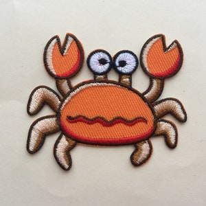 May include: A cartoon crab with orange and brown embroidery. The crab has large white eyes and a red mouth.