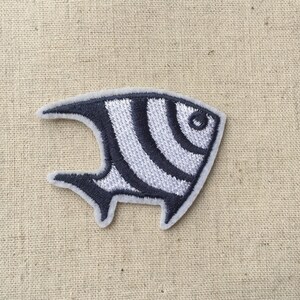 Può includere: Una toppa da stirare ricamata in bianco e nero di un pesce stilizzato con un corpo curvo e un solo occhio.