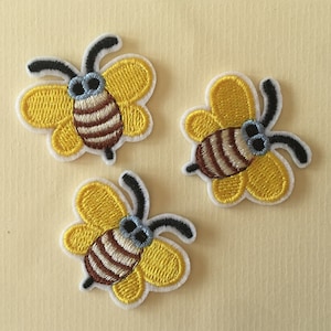 May include: Three yellow and brown embroidered bee patches with white outlines. The bees have black antennae and blue eyes.