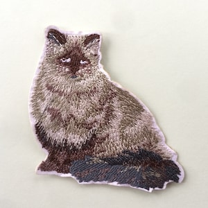 Peut inclure: Patch brodé représentant un chat brun et blanc assis, la queue enroulée autour de ses pattes.