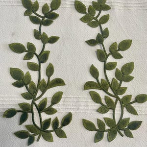 May include: Two embroidered green leaf branch appliques. Each applique features a central stem with multiple leaves. The leaves are a darker shade of green, and the background is a white, textured fabric.