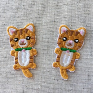 May include: Two embroidered iron-on patches of orange tabby cats wearing green collars. The cats are facing forward with their mouths closed and their eyes closed.