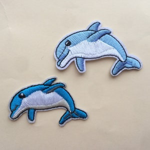 May include: Two embroidered blue and white dolphin patches. The dolphins are facing the right side of the image and are jumping out of the water.