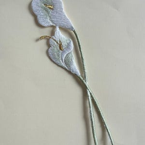 May include: Embroidered patch featuring two white calla lilies with gold accents. The stems are a light green color. The patch is designed to be sewn onto fabric, adding a floral touch to clothing or accessories.