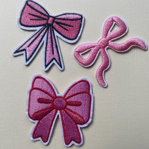 Könnte beinhalten: Drei gestickte Aufnäher mit rosa Schleifen. Die Schleifen haben unterschiedliche Designs, eine mit schwarzen Details und eine andere mit einer fließenden Form. Alle sind weiß umrandet, vor einem neutralen Hintergrund.