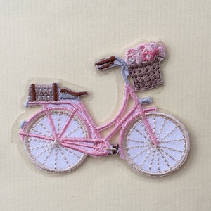 Op de afbeelding: Een roze fiets met een bruine mand gevuld met roze bloemen. De fiets heeft een bruin zadel en een bruine mand achterop.