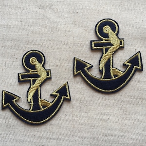 May include: Two navy blue anchor patches with gold embroidered details. The anchors have a classic design, featuring a rope wrapped around the central post. The patches are likely for sewing onto clothing or accessories.