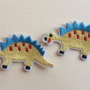 May include: Two yellow embroidered dinosaur patches with blue spines and red spots. The dinosaurs have black eyes and black feet.