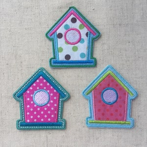 Op de afbeelding: Drie geborduurde vilt vogelhuisjes. De vogelhuisjes zijn roze, blauw en groen met witte stippen en een lichtblauwe cirkel voor het raam.