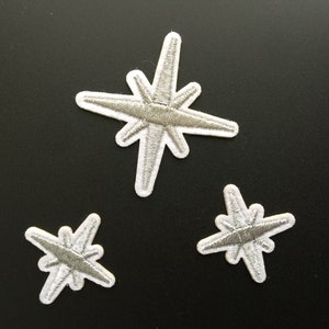 以下が含まれることがあります： さまざまなサイズの刺繍された星のパッチが3つ。星は白の縁取りが付いたシルバーで、黒い背景に配置されています。これらのパッチは、衣類やアクセサリーを飾るために使用できます。