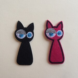 Könnte beinhalten: Zwei Katzen-Applikationen aus Filz. Eine ist schwarz, die andere rosa. Beide haben gestickte Details: blaue Augen, graue Augenlider und eine weiße Kontur. Die Katzen-Applikationen sind stilisiert und skurril.