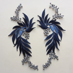 May include: Two blue and silver floral appliques with a leafy design. The appliques are made of fabric and have a delicate, feminine look.