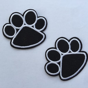 Puede incluir: Dos parches de huella de pata negra con contornos blancos. Las huellas de pata están bordadas y tienen una tela texturizada.