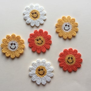 Puede incluir: Seis aplicaciones de flores de ganchillo con caras sonrientes. Tres son amarillas, dos son naranjas y una es blanca.