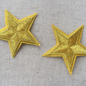 May include: Two gold star-shaped fabric patches. The stars are embroidered with gold thread and have a five-pointed design.