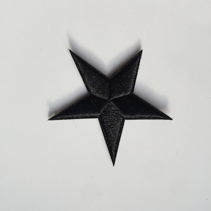 May include: A black, five-pointed star patch on a white background. The patch has a textured surface, with each point clearly defined. The star's design is simple and bold, suitable for various applications. The patch is likely for clothing or accessories.