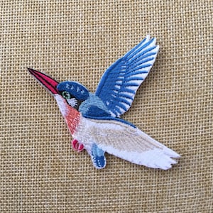 Pode incluir: Um remendo bordado azul e branco para passar a ferro com a imagem de um guarda-rios em voo. O pássaro tem um bico longo e fino e um corpo azul brilhante com asas brancas.