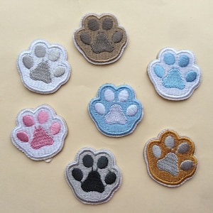 Könnte beinhalten: Sechs gestickte Bügelbilder in Form einer Pfote. Die Patches sind weiß mit verschiedenen farbigen Pfotenabdrücken in der Mitte. Die Farben sind grau, hellblau, rosa, dunkelblau, hellbraun und gold.