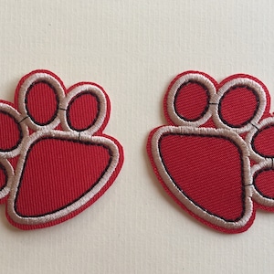 May include: Two red paw print patches with tan and black embroidered details. The patches are embroidered with a red fabric and have a tan border and black outline. The patches are likely for pets.