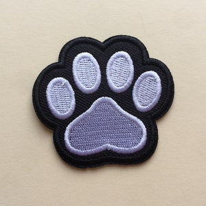 May include: Embroidered paw print patch in black with light purple details. The patch features a black border and a detailed paw pad and toe pads in light purple. This patch is ideal for adding to clothing, bags, or other fabric items.