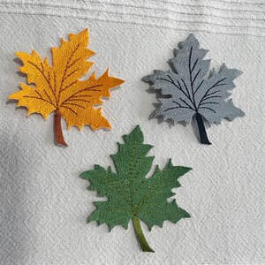 May include: Three embroidered maple leaf patches in autumnal colours. One is golden yellow with brown veins and stem, another is grey with black veins and stem, and the third is green with a green stem. The leaves are arranged on a white textured surface.