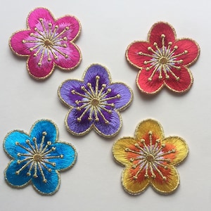 Puede incluir: Cinco parches de flores bordadas en diferentes colores. Las flores son rosa, roja, morada, azul y naranja. Cada flor tiene un centro dorado y un diseño de cinco pétalos.