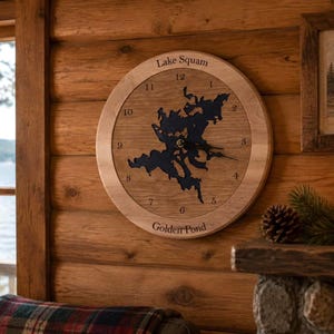 May include: A round wooden wall clock featuring a dark blue cutout map of Lake Squam in the center. The clock face displays black numbers from 1 to 12, with the text Lake Squam at the top and Golden Pond at the bottom in black lettering.
