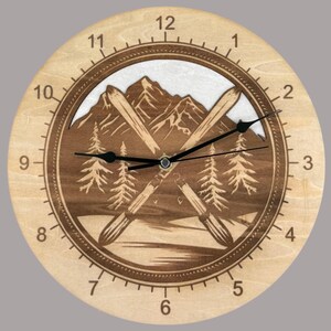 May include: A round wooden clock with a mountain and ski design. The clock face features a mountain range, crossed skis, and trees. The clock hands are black, and the numbers are etched around the edge. The clock is approximately 25 cm in diameter.