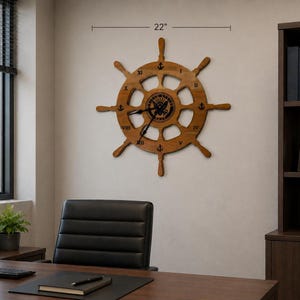 Puede incluir: Un reloj de pared de madera con forma de timón de barco de 55,88 cm con acabado marrón natural, números romanos negros e iconos de anclas negras en cada hora. El centro muestra el emblema negro de la Marina de los EE. UU. con manecillas negras.