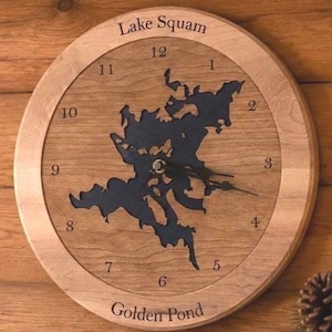 Puede incluir: Un reloj de pared redondo de madera con un mapa recortado en azul oscuro del lago Squam en el centro. La esfera muestra números negros del 1 al 12. El texto Lake Squam está grabado en la parte superior y Golden Pond en la parte inferior.