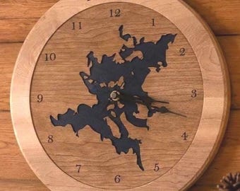 May include: A round wooden wall clock featuring a dark blue cutout map of Lake Squam in the center. The clock face displays black numerals from 1 to 12. The text Lake Squam is engraved at the top and Golden Pond is engraved at the bottom.