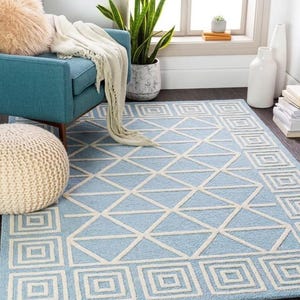 May include: A rectangular area rug featuring a light blue background with a white geometric pattern consisting of interconnected triangles and a border of concentric squares. The rug has a textured pile surface and covers a dark wooden floor area.