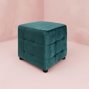 Puede incluir: Un otomán cúbico de color verde azulado con un diseño acolchado. La tela afelpada, similar al terciopelo, está dividida en secciones cuadradas. El otomán se apoya sobre cuatro pequeños pies negros, ofreciendo una elegante opción de asiento. El fondo es rosa pálido.