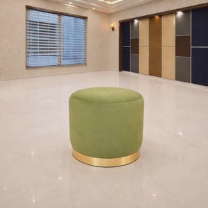 Pode incluir: Um pufe redondo verde-oliva com uma base dourada. O pufe é estofado em tecido macio e aveludado e assenta numa faixa metálica brilhante. A sala tem uma janela grande e uma parede decorativa.