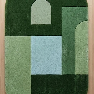 May include: A rectangular rug with rounded corners, featuring a geometric design in shades of green and a light blue square. The rug's design includes arch and rectangular shapes, creating a modern, abstract pattern. The rug is made of a soft, plush material.