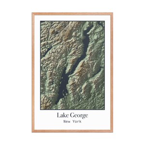 May include: A framed topographical map of Lake George, New York, in shades of green, brown, and blue. The map is encased in a light brown wooden frame, with the lake's name and state printed in a white font at the bottom.