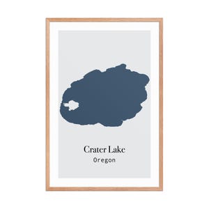 Może przedstawiać: Oprawiony wydruk z minimalistyczną ilustracją Crater Lake w Oregonie. Jezioro przedstawione jest w ciemnoniebieskim odcieniu na jasnoszarym tle. Tekst "Crater Lake Oregon" jest wyśrodkowany pod ilustracją jeziora.