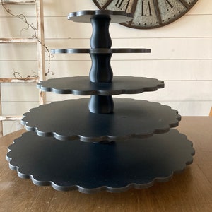 May include: A black, four-tiered serving stand with scalloped edges. The stand has a central support column and is displayed on a wooden table. The background includes a large clock and a white wall.