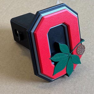 May include: A black trailer hitch cover featuring a red "O" logo with a green leaf and a brown nut. The logo is outlined in gray and black. The hitch cover is a sports-themed accessory.